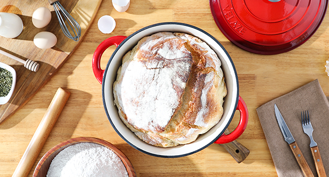 Pain fraîchement cuit dans un four hollandais rouge, entouré d'ingrédients de cuisson et d'ustensiles sur une surface en bois.