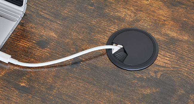 Passe-fil de bureau circulaire traversé par un câble blanc, installé sur une surface en bois à côté d'un appareil électronique partiellement visible