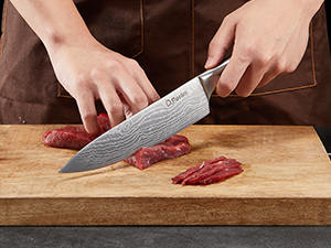 pour trancher de la viande crue sur une planche à découper en bois. Mains visibles tenant le couteau et coupant de fines tranches de viande rouge.