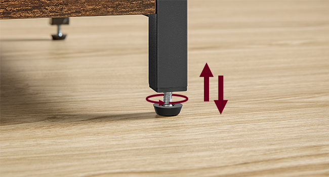 Gros plan d'un ajusteur de pieds de meuble sur un plancher en bois. Base cylindrique noire avec cadran circulaire rouge pour le réglage de la hauteur, indiqué par des flèches vers le haut et vers le bas.