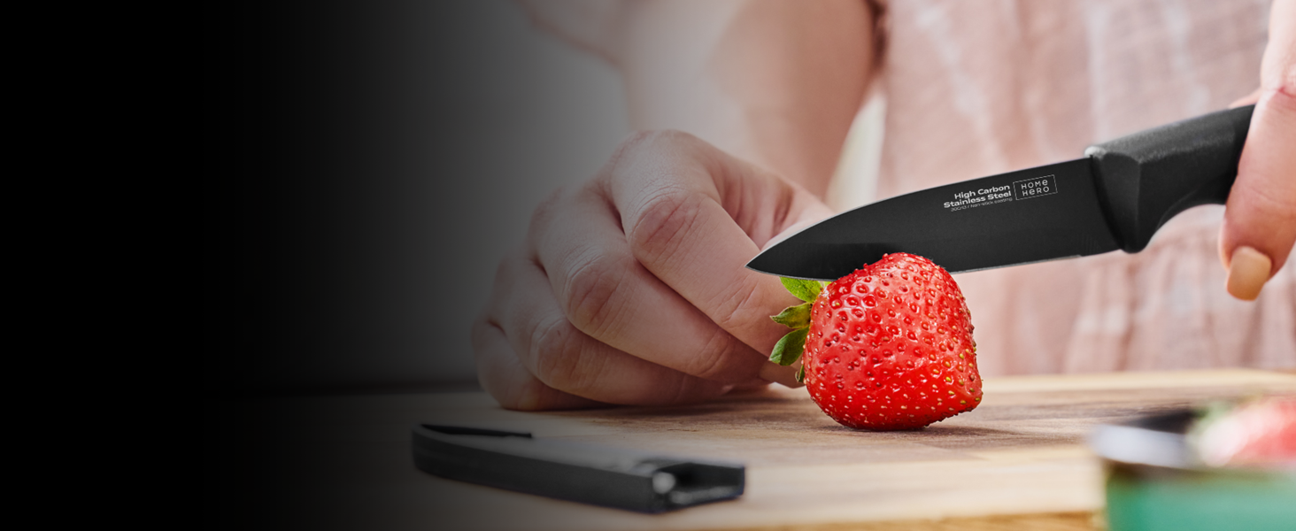 Tenez à la main un couteau de cuisine noir et coupez une fraise rouge bien mûre sur une planche à découper en bois. Un smartphone est visible en arrière-plan.