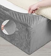 Gros plan d'une main soulevant le coin d'un drap-housse gris, révélant son bord élastique et son ajustement parfait sur un matelas