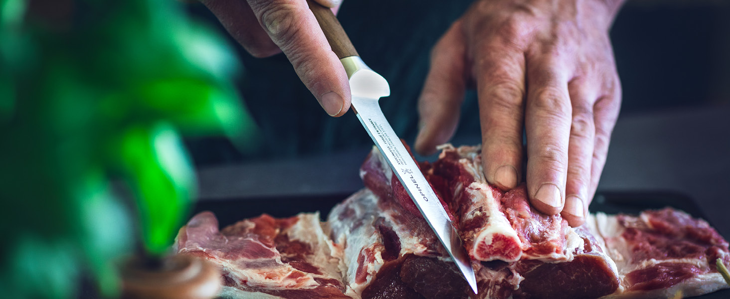 Gros plan des mains à l'aide d'un couteau bien aiguisé pour couper de la viande crue sur une surface sombre, avec un feuillage vert visible en arrière-plan