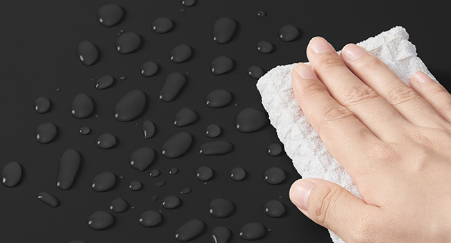 Gros plan d'une main essuyant les gouttelettes d'eau d'une surface sombre et résistante à l'eau avec un chiffon blanc, démontrant ainsi ses propriétés imperméables.