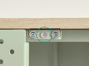 Gros plan d'un mécanisme de joint de meuble. Un support métallique à ouvertures circulaires relie un plateau en bois de couleur claire à un cadre ou à un panneau en métal vert pâle