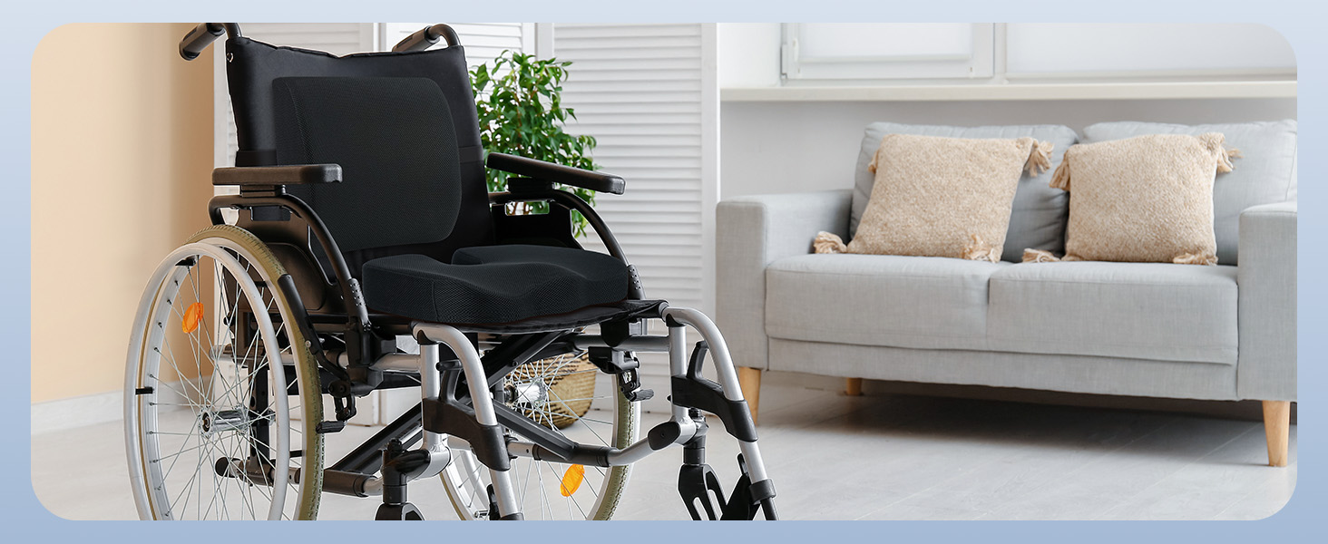 Fauteuil roulant manuel noir avec structure métallique et grandes roues, positionné au premier plan. Canapé gris clair avec pieds en bois et coussins beiges visibles en arrière-plan.