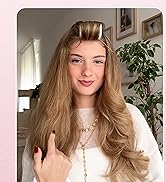 Cheveux blonds longs et volumineux coiffés avec une partie avant relevée. Pull en maille blanche et délicat collier doré visible. Fond intérieur avec plante et œuvres d'art encadrées.