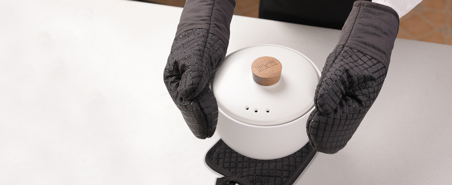 Des mains portant des gants de cuisine gris tiennent une marmite en céramique blanche avec un couvercle en liège, démontrant ainsi la fonctionnalité de protection thermique