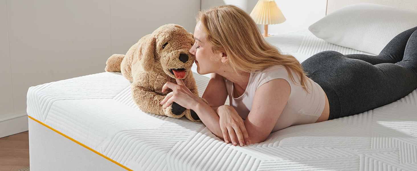 Matelas avec bordure jaune sur plancher en bois. Femme allongée sur un matelas, interagissant avec un ours en peluche. Literie blanche visible.
