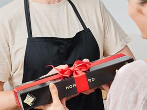 Personne en tablier noir tenant une boîte-cadeau foncée avec le texte « Homero » et le logo en feuille d'or. La boîte a un ruban rouge et un nœud. Les mains d'une autre personne sont visibles en train de recevoir le cadeau.