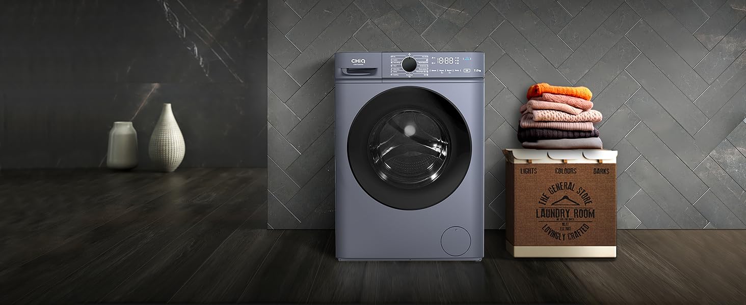 avec extérieur gris et porte vitrée circulaire. Panneau de commande numérique sur le dessus. Caisse en bois avec vêtements pliés à proximité.