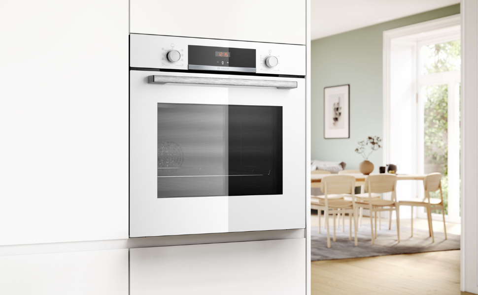 Four mural de cuisine blanc encastré présenté dans un décor de cuisine moderne avec coin repas visible en arrière-plan. Le design élégant comprend une façade en verre et un panneau d'affichage numérique.