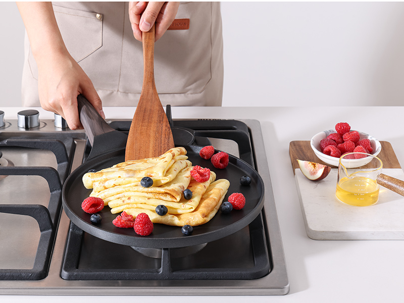 Scène de cuisine avec une crêpe ou une fine crêpe préparée dans une poêle noire, garnie de framboises et de myrtilles fraîches