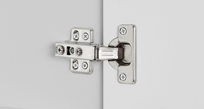 Gros plan d'un mécanisme de charnière d'armoire au fini argenté/métallique, montrant la plaque de montage et le bras fixés à la porte et au cadre de l'armoire.