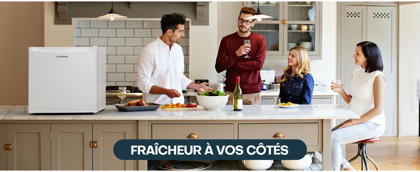Réfrigérateur compact blanc dans une cuisine moderne avec des gens rassemblés autour de l'île. Le texte superposé indique « FRAÎCHEUR À VOS CÔTÉS
