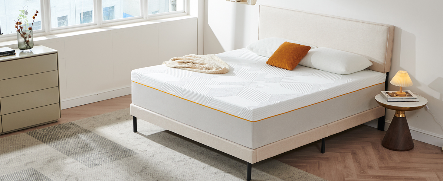 Scène de chambre à coucher avec un matelas blanc sur un cadre de lit de couleur claire. Coussin orange et couverture pliée visibles. Table d'appoint avec lampe et commode en arrière-plan.